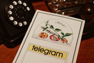 Na drewnianym, brązowym biurku leży biały blankiet telegramowy. W jego górnej części grafika wielkanocna, dwie kolorowe pisanki, bazie i napis 'Alleluja', w dolnej części czarny napis 'telegram' na żółtym tle i puste miejsce na wiadomość telegramu. W lewym górnym rogu fotografii fragment tarczy starego, czarnego telefonu.