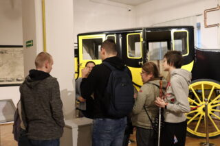 Grupa 4 osób zwróconych tyłem do aparatu. Oglądają niewidoczny na zdjęćiu eksponat muzealny. W tle przewodnik oraz czarno- żółty dyliżans pocztowy.