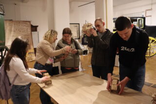 Zdjęcie pięciu osób, które stoją przy jasnym muzealnym stole i poznają przez dotyk drewniane miniatury dyliżansu pocztowego.