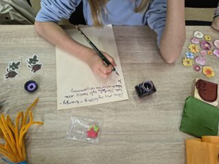 Zbliżenie na dłoń dziecka używającą pióra i atramentu do pisania listu lub ozdabiania kartki, obok widoczny kałamarz.