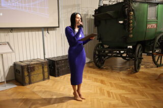Dyrektor Muzeum w niebieskiej sukni stojąca przed wystawą i wygłaszająca przemówienie podczas wernisażu.