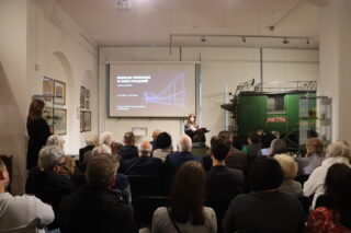 Szeroki kadr przedstawiający salę muzealną wypełnioną publicznością, która siedzi w rzędach i słucha prelekcji. W centrum widoczny jest duży ekran projekcyjny z tytułem wystawy 'Breslau-Wrocław w sieci połączeń' i schematycznym rysunkiem mostu. Obok ekranu stoi prelegentka, a na prawym planie widoczny jest fragment dużego, zielonego furgonu pocztowego