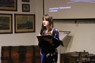 Kuratorka wystawy, ubrana w ciemną sukienkę z białym lub jasnym wzorem, stoi przed ekranem projekcyjnym i trzyma mikrofon oraz czarną teczkę, wygłaszając przemówienie. W tle widoczne są stare, drewniane kufry oraz obrazy w ramkach na ścianie. Na ekranie widnieje napis: 'Muzeum Poczty i Telekomunikacji we Wrocławiu'.