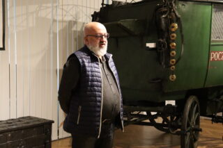 Michał Grzesik przemawiający podczas wernisażu. Znajduje się obok dużego, historycznego, zielonego furgonu pocztowego.