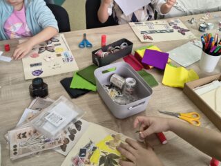 Dłoń dziecka w trakcie klejenia ozdób do kartki; w tle koszyki i pojemniki wypełnione pomponami i koralikami.