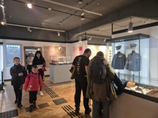 Rodziny zwiedzają salę wystawową z umundurowaniem i eksponatami pocztowymi.