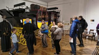 Grupa osób (zwiedzających) stoi na drewnianej podłodze w muzeum i ogląda duży, historyczny, czarno-żółty dyliżans pocztowy, widoczny w tle. Dyliżans ma czerwone detale i bagaż na dachu.