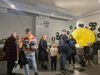 Zwiedzający stoją przy instalacji z żółtym megafonem i planszą o historii łączności.